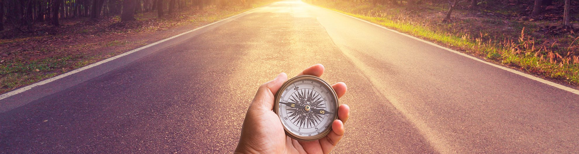 Hand holding compass on empty asphalt road and sunset.